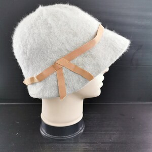 May include: A light gray cloche hat with a soft, fuzzy texture. The hat features a small brim and is adorned with a tan ribbon tied in a bow. The hat is displayed on a mannequin head.