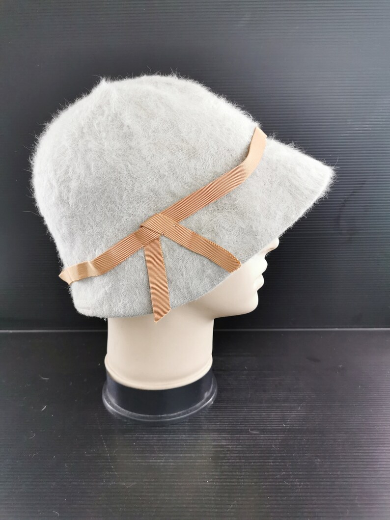 May include: A light gray cloche hat with a soft, fuzzy texture. The hat features a small brim and is adorned with a tan ribbon tied in a bow. The hat is displayed on a mannequin head.