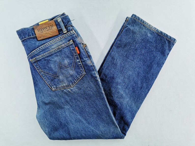 May include: Blue denim jeans with a classic five-pocket design. Features a brown leather patch with "EDWIN US CLASSIC" branding and a red logo tag. The jeans have a straight leg cut.