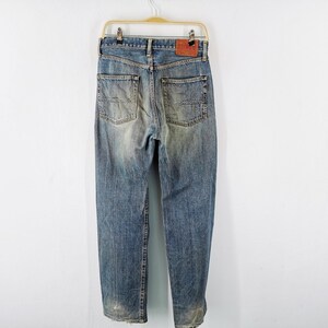 May include: Faded blue denim jeans with a worn appearance, hanging on a gold-colored hanger. The jeans have a classic five-pocket design, a leather patch, and a straight leg cut. The denim has a distressed look.