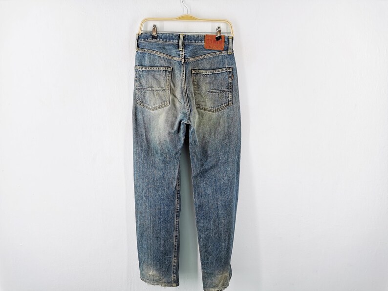 May include: Faded blue denim jeans with a worn appearance, hanging on a gold-colored hanger. The jeans have a classic five-pocket design, a leather patch, and a straight leg cut. The denim has a distressed look.