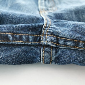 May include: Close-up of blue denim jeans with visible stitching in a contrasting tan color. The image highlights the seam details and texture of the fabric, showcasing the construction of the garment.