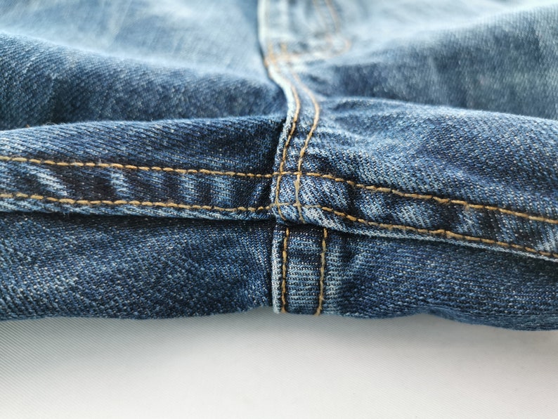 May include: Close-up of blue denim jeans with visible stitching in a contrasting tan color. The image highlights the seam details and texture of the fabric, showcasing the construction of the garment.