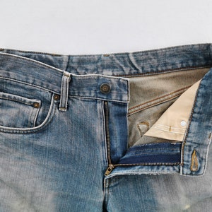 May include: Close-up of blue denim jeans with a partially unzipped fly. The jeans have a classic five-pocket design, with visible stitching and metal rivets. The denim has a faded, worn appearance.