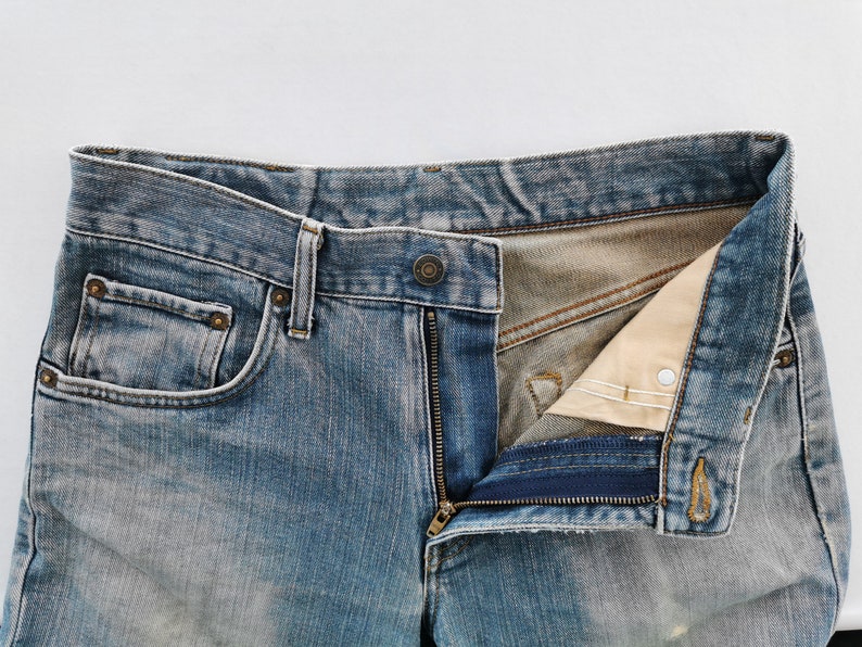 May include: Close-up of blue denim jeans with a partially unzipped fly. The jeans have a classic five-pocket design, with visible stitching and metal rivets. The denim has a faded, worn appearance.