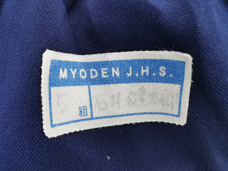 Puede incluir: Primer plano de una tela azul marino con una etiqueta blanca. La etiqueta dice "MYODEN J.H.S." en azul. Debajo, dos cuadros en blanco y un cuadro m&aacute;s peque&ntilde;o con caracteres japoneses.