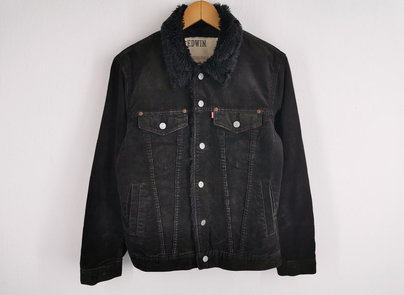 Puede incluir: Chaqueta de pana negra con cuello de piel sint&eacute;tica. La chaqueta tiene dos bolsillos en el pecho y dos bolsillos laterales, con botones plateados y una etiqueta roja. La chaqueta es de la marca Edwin.