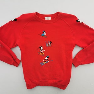 Puede incluir: Sudadera roja con Mickey Mouse esquiando. Las mangas tienen la palabra "MOUSE" en letras blancas. Mickey Mouse se representa en varias poses de esquí en la parte delantera de la sudadera.