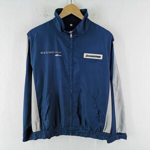Puede incluir: Chaqueta azul marino con cremallera y mangas y detalles grises. La chaqueta presenta el texto "REGNO GR-XT" y un logotipo de "BRIDGESTONE". Tiene cuello alzado y dos bolsillos delanteros. La chaqueta está colgada en una percha de madera.