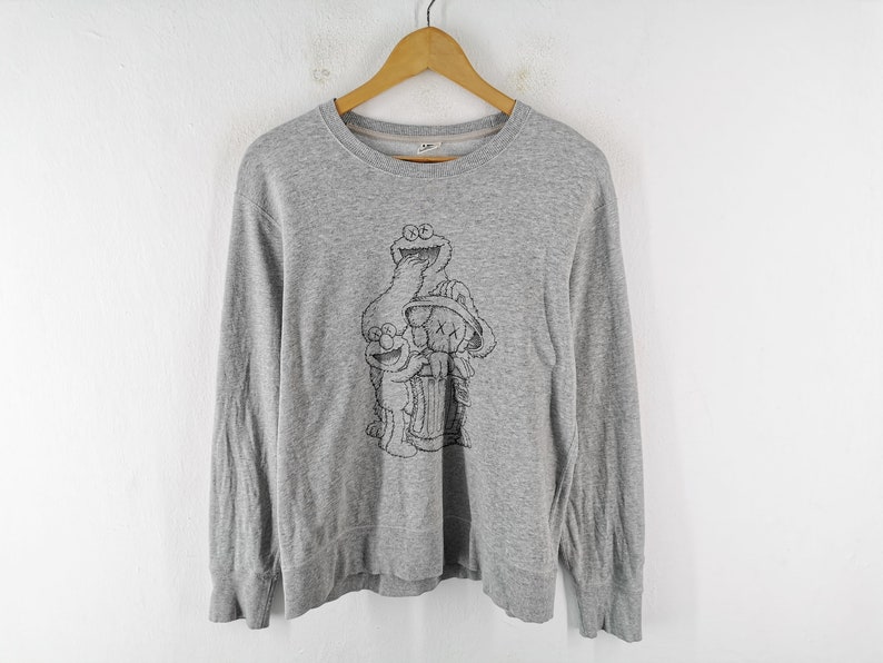 K&ouml;nnte beinhalten: Ein graues Langarm-Sweatshirt mit einer schwarz-wei&szlig;en Grafik von Sesamstra&szlig;en-Charakteren. Das Design umfasst Kr&uuml;melmonster, Elmo und eine Figur mit durchgestrichenen Augen. Das Sweatshirt h&auml;ngt an einem Holzkleiderb&uuml;gel.