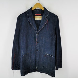 May include: A dark blue denim blazer with a three-button closure, two front pockets, and red stitching details. The jacket is hanging on a wooden hanger against a white background. A tag is attached to the jacket.