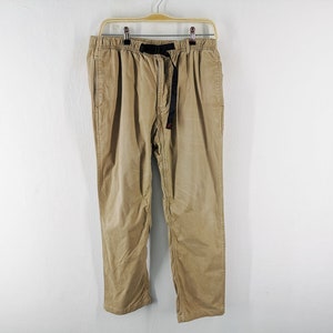 May include: Tan corduroy pants hanging on a wooden hanger against a white wall. The pants have a black belt with a red accent. The pants have a relaxed fit and a drawstring waist.