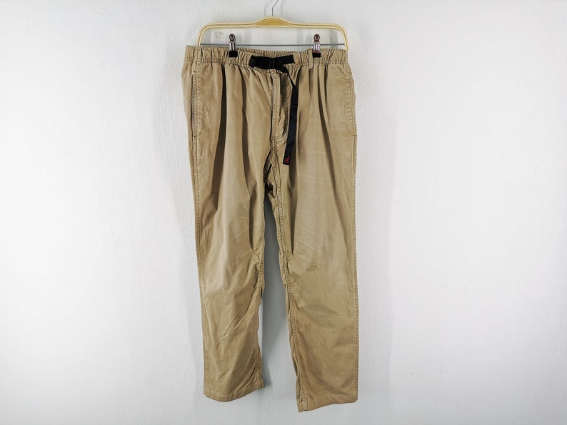 May include: Tan corduroy pants hanging on a wooden hanger against a white wall. The pants have a black belt with a red accent. The pants have a relaxed fit and a drawstring waist.