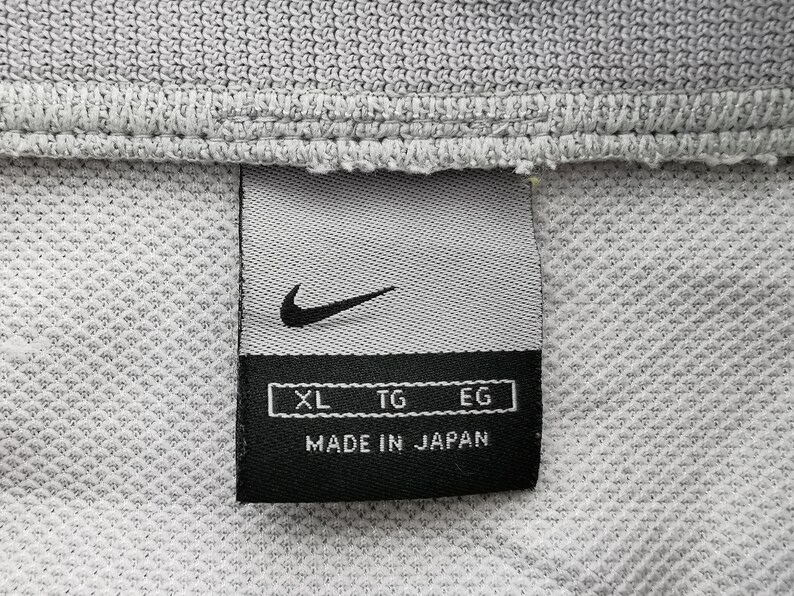 Puede incluir: Primer plano de una tela gris con una etiqueta negra de Nike. La etiqueta presenta el logotipo de Nike, indicadores de talla XL, TG, EG y el texto "MADE IN JAPAN". La tela tiene un patr&oacute;n texturizado en forma de rejilla.