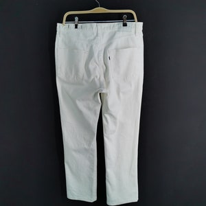 May include: Cream-coloured denim trousers hanging on a wooden hanger against a dark grey background. The trousers have two back pockets and a small label. The trousers are straight-legged.