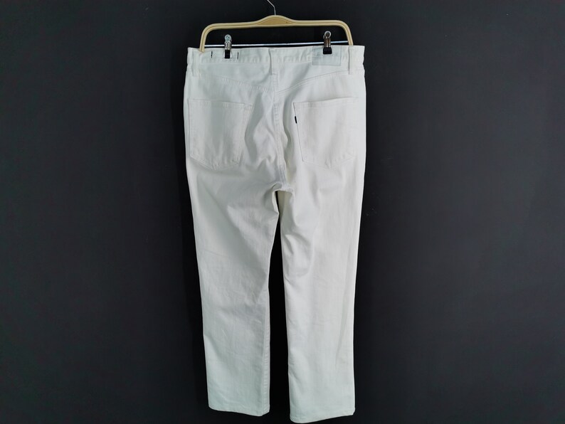 May include: Cream-coloured denim trousers hanging on a wooden hanger against a dark grey background. The trousers have two back pockets and a small label. The trousers are straight-legged.