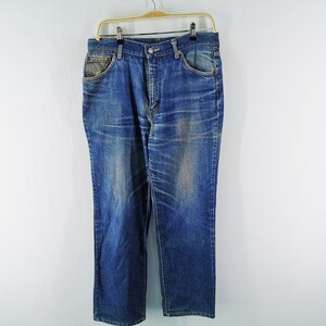 May include: Blue denim jeans hanging on a gold hanger. The jeans have a classic five-pocket design with a button and zipper closure. The denim has a faded, worn look with lighter areas on the thighs and knees.