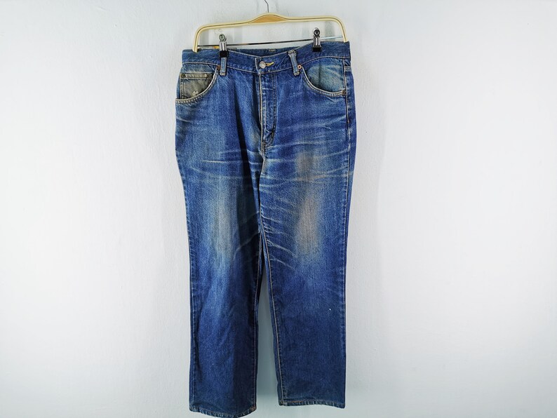 May include: Blue denim jeans hanging on a gold hanger. The jeans have a classic five-pocket design with a button and zipper closure. The denim has a faded, worn look with lighter areas on the thighs and knees.