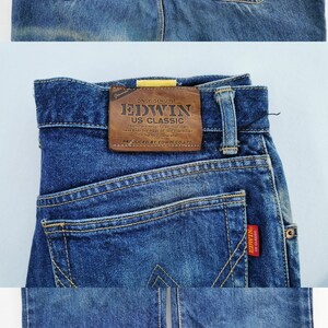 May include: Close-up of blue denim jeans with a classic five-pocket design. Features a brown leather patch with "EDWIN US CLASSIC" branding. The back pockets have a distinctive "W" stitch. A red "EDWIN" tag is visible.