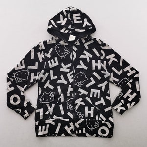 May include: Black zip-up hoodie with a repeating pattern of white letters and Hello Kitty faces. The hoodie has a drawstring hood and a full-length zip. The design is printed on a black background.