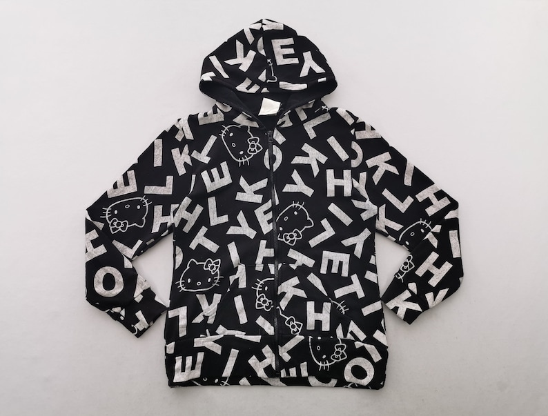 May include: Black zip-up hoodie with a repeating pattern of white letters and Hello Kitty faces. The hoodie has a drawstring hood and a full-length zip. The design is printed on a black background.