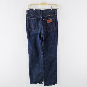 Op de afbeelding: Donkerblauwe denim jeans hangend aan een hanger. De jeans hebben een klassiek ontwerp met vijf zakken en een bruine leren patch op de achterzak. Het Wrangler-logo is zichtbaar op de patch. De jeans hebben een rechte pijp.