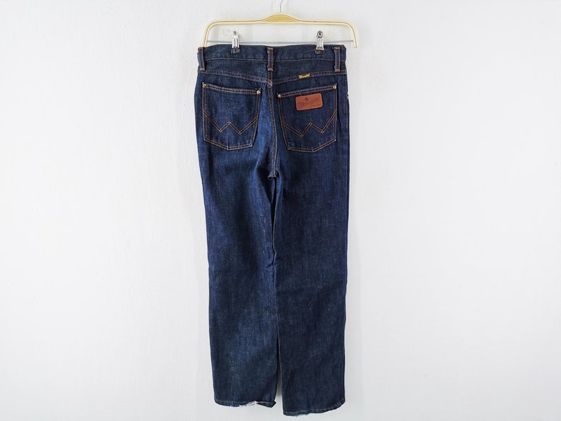 Op de afbeelding: Donkerblauwe denim jeans hangend aan een hanger. De jeans hebben een klassiek ontwerp met vijf zakken en een bruine leren patch op de achterzak. Het Wrangler-logo is zichtbaar op de patch. De jeans hebben een rechte pijp.