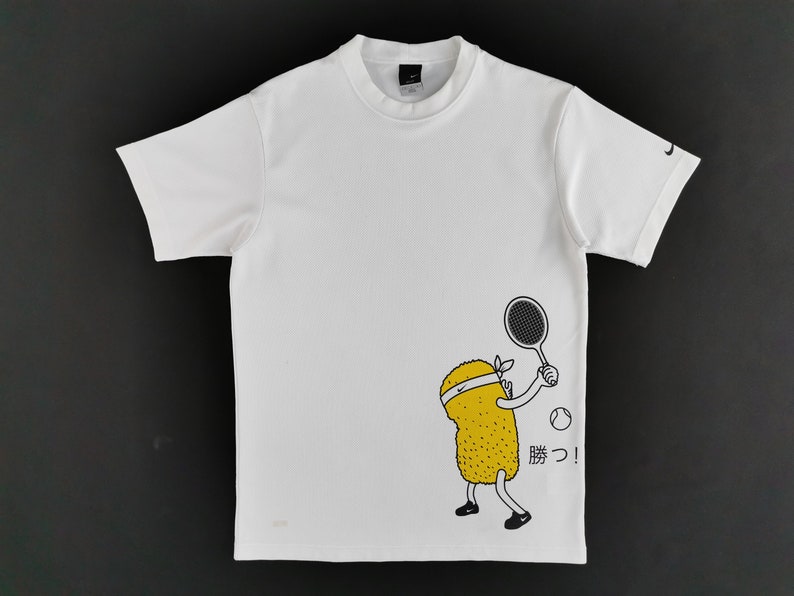 May include: White short-sleeved t-shirt featuring a cartoon tennis player. The graphic shows a yellow character with a headband, holding a tennis racket and a tennis ball. The Japanese word "勝つ!" is printed next to the character.