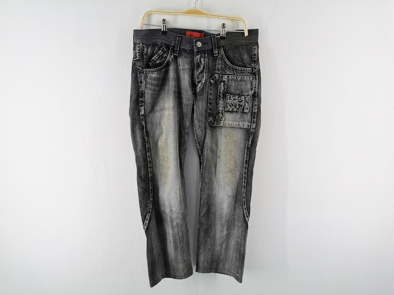May include: Faded black denim jeans with unique pocket details. Features a patch pocket on the right leg and standard pockets. The jeans are hanging on a wooden hanger against a white background.