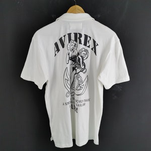 Puede incluir: Polo blanco con un gr&aacute;fico negro en la espalda. El gr&aacute;fico incluye la palabra "AVIREX", una pin-up y un ancla. El texto "A smooth sea never made a skilled sailor" est&aacute; debajo.