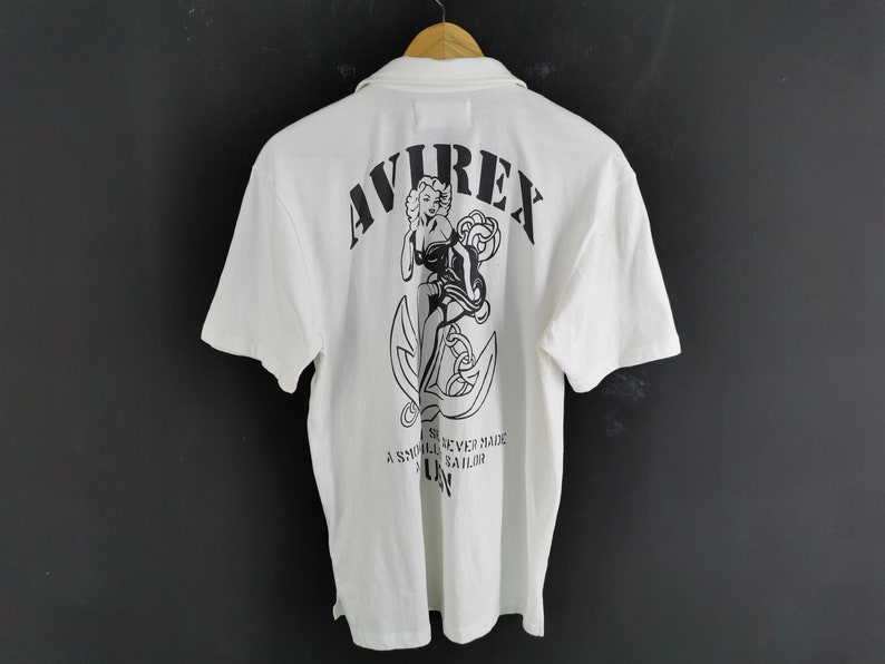 Puede incluir: Polo blanco con un gr&aacute;fico negro en la espalda. El gr&aacute;fico incluye la palabra "AVIREX", una pin-up y un ancla. El texto "A smooth sea never made a skilled sailor" est&aacute; debajo.
