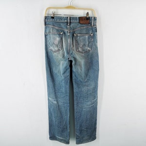 May include: Light blue denim jeans with a faded wash, hanging on a wooden hanger. The jeans have a classic five-pocket design, a leather patch with the brand name, and a straight leg fit.