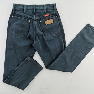 K&ouml;nnte beinhalten: Dunkelblaue Jeans aus Denim mit klassischem F&uuml;nf-Taschen-Design. Mit rotem Wrangler-Label und braunem Lederaufn&auml;her. Die Jeans haben einen geraden Beinverlauf und liegen flach auf einer wei&szlig;en Oberfl&auml;che.