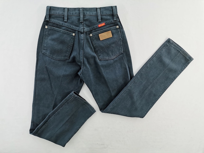 K&ouml;nnte beinhalten: Dunkelblaue Jeans aus Denim mit klassischem F&uuml;nf-Taschen-Design. Mit rotem Wrangler-Label und braunem Lederaufn&auml;her. Die Jeans haben einen geraden Beinverlauf und liegen flach auf einer wei&szlig;en Oberfl&auml;che.