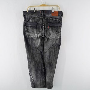 May include: Dark gray denim jeans with a faded wash, featuring two back pockets and a small red tag. The jeans are hanging on a wooden hanger against a white background. A brown leather patch is visible.