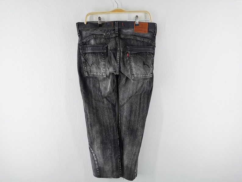 May include: Dark gray denim jeans with a faded wash, featuring two back pockets and a small red tag. The jeans are hanging on a wooden hanger against a white background. A brown leather patch is visible.