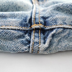Op de afbeelding: Close-up van een blauwe denim jeans, met de nadruk op de stiksels en de textuur van de stof. De afbeelding toont de details van de naden en de algehele constructie van het kledingstuk. De denim heeft een vervaagde uitstraling.