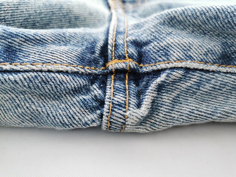 Op de afbeelding: Close-up van een blauwe denim jeans, met de nadruk op de stiksels en de textuur van de stof. De afbeelding toont de details van de naden en de algehele constructie van het kledingstuk. De denim heeft een vervaagde uitstraling.