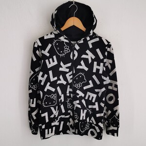 May include: Black zip-up hoodie featuring a pattern of white letters and Hello Kitty faces. The hoodie has a black hood and is suspended on a wooden hanger. The design covers the front and sleeves.