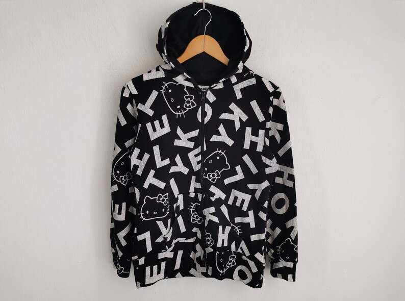 May include: Black zip-up hoodie featuring a pattern of white letters and Hello Kitty faces. The hoodie has a black hood and is suspended on a wooden hanger. The design covers the front and sleeves.