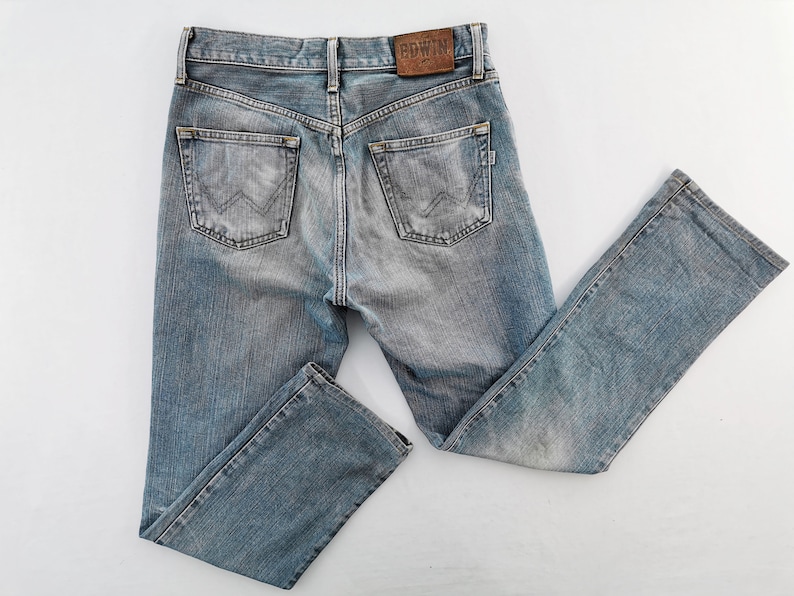 May include: Light blue denim jeans with a faded wash. The jeans have a classic five-pocket design, a straight leg, and a leather patch with the brand name "EDWIN" on the back. The jeans are laid flat on a white surface.