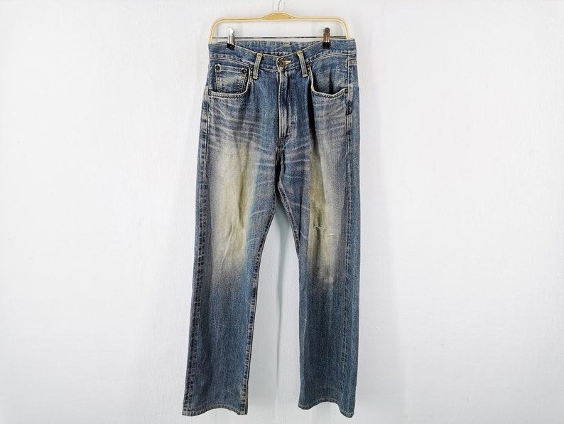 May include: Blue denim jeans with a faded wash and distressed details, hanging on a wooden hanger. The jeans have a classic five-pocket design and a straight leg fit. The fabric shows signs of wear.