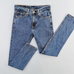 May include: Blue denim jeans with a classic five-pocket design. The jeans have a button and zipper closure, belt loops, and copper-colored rivets. The fabric has a textured appearance, and the jeans are laid flat on a white surface.