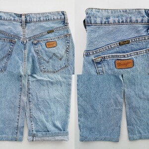 May include: Light blue denim Wrangler shorts. The shorts have a classic five-pocket design, a brown leather Wrangler patch, and a Wrangler label on the back pocket. The shorts have a cuffed hem.