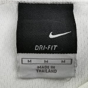 May include: Close-up of a white athletic shirt label. The black label features the Nike swoosh logo and the words "DRI-FIT". Below are three "M" size labels and the text "MADE IN THAILAND".