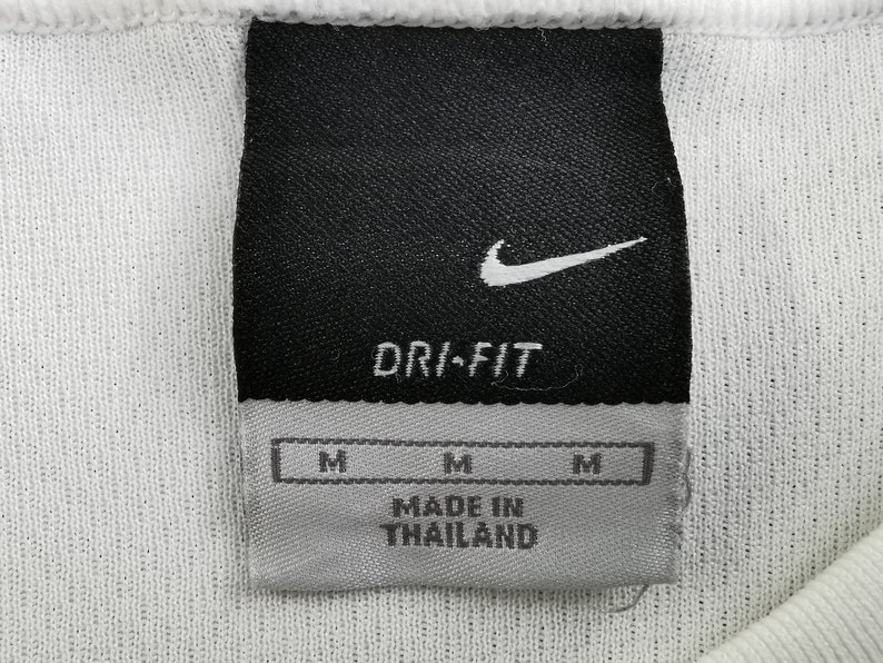 May include: Close-up of a white athletic shirt label. The black label features the Nike swoosh logo and the words "DRI-FIT". Below are three "M" size labels and the text "MADE IN THAILAND".