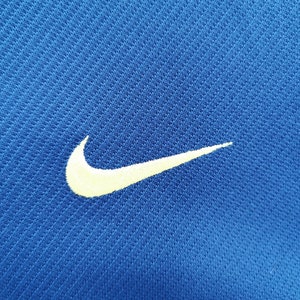 May include: Close-up of a blue athletic shirt with a white Nike swoosh logo embroidered on the left side. The fabric has a textured, breathable appearance.