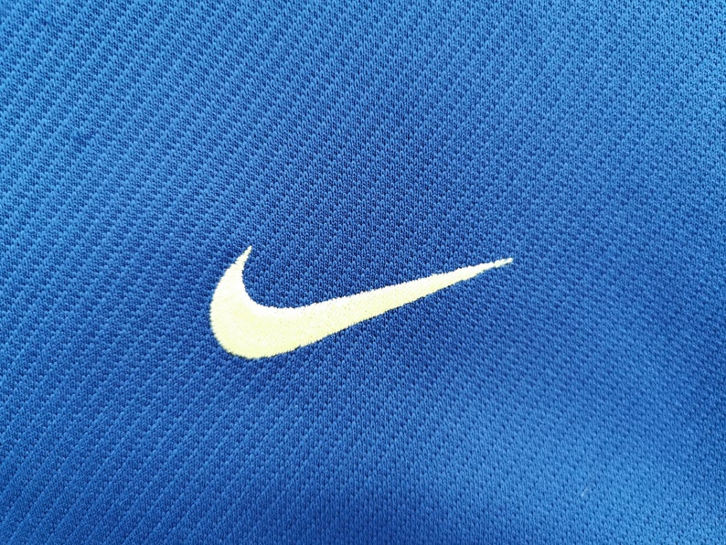 May include: Close-up of a blue athletic shirt with a white Nike swoosh logo embroidered on the left side. The fabric has a textured, breathable appearance.