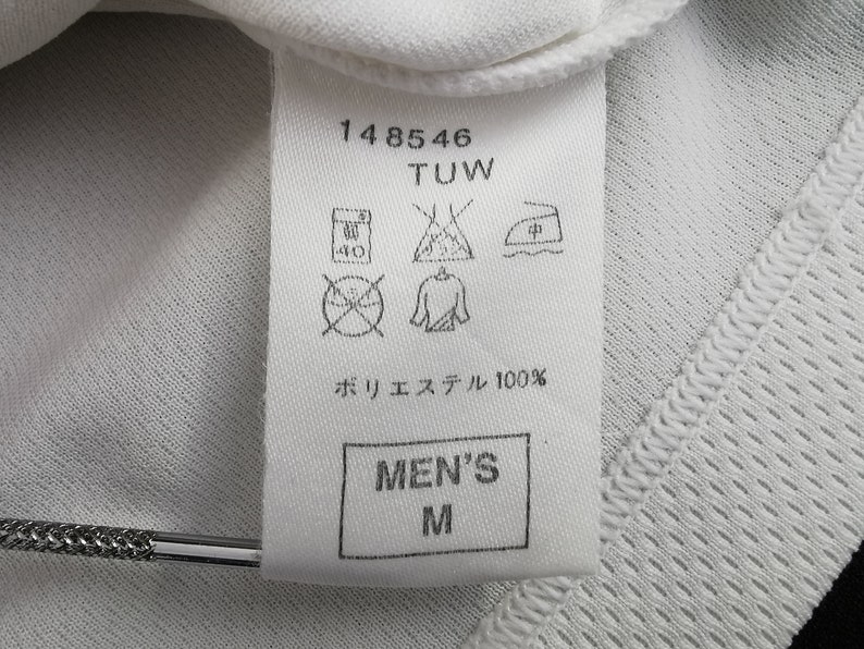May include: Close-up of a white garment label with the text "148546 TUW". The label features care symbols and the text "ポリエステル 100%" and "MEN'S M". The garment has a textured, breathable fabric.