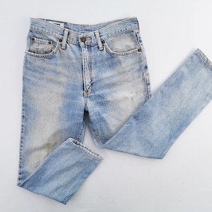 Puede incluir: Vaqueros de mezclilla azul claro con un lavado descolorido y un dise&ntilde;o cl&aacute;sico de cinco bolsillos. Los vaqueros tienen cierre de bot&oacute;n y cremallera, trabillas para el cintur&oacute;n y un aspecto ligeramente desgastado. Los vaqueros est&aacute;n colocados sobre una superficie blanca.